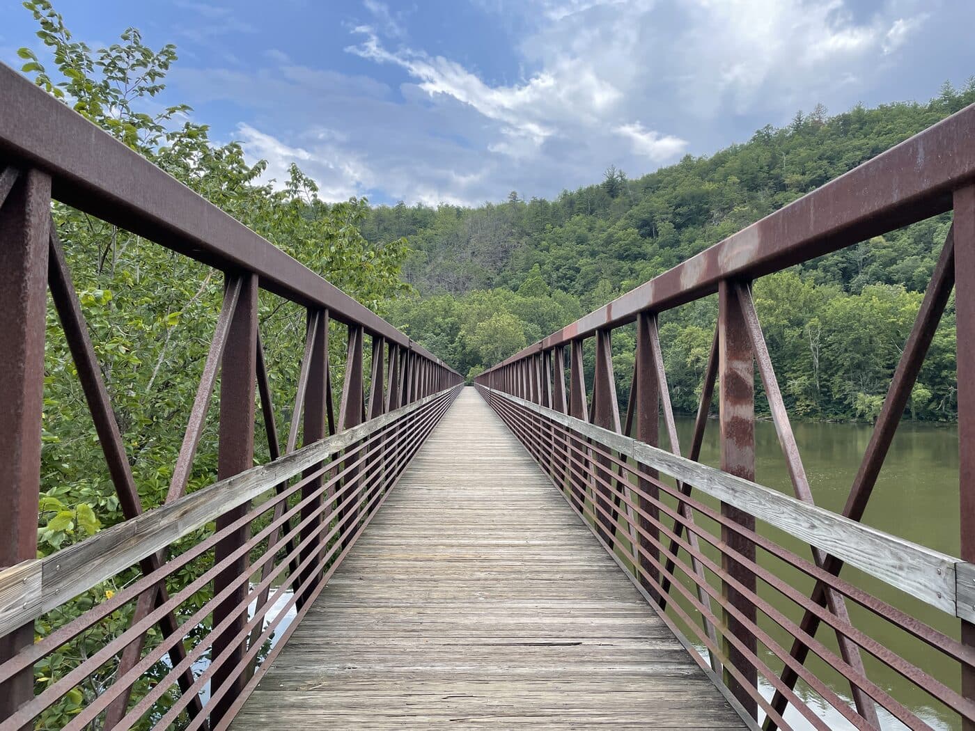 Looking along the deck of the James River Footbridge on the Appalachian Trail, rusted steel trusses converging toward the wooded far bank under a cloudy summer sky.