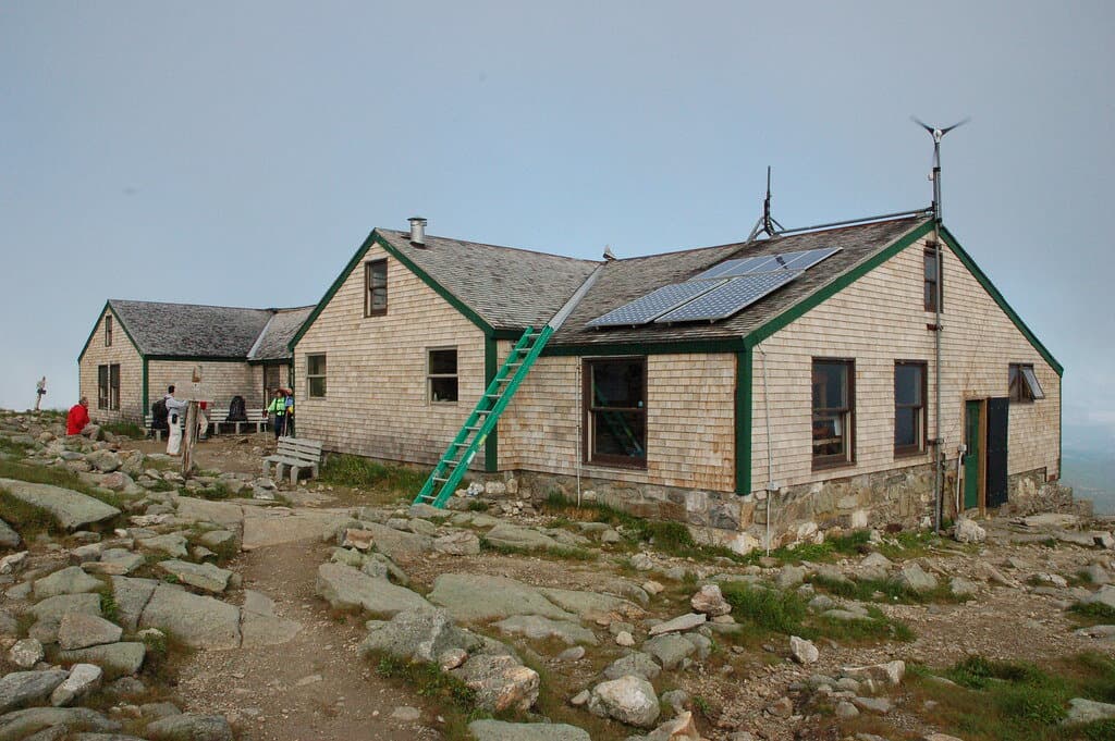 Lakes of the Clouds Hut, a long shingled building with green trim and rooftop solar panels, on the rocky alpine shoulder of Mount Washington with hikers resting outside.